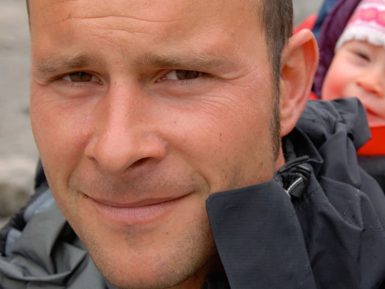 Bergführer Uwe Berg lächelt in grauer Funktionsjacke, neben ihm eine Person mit bunter Wintermütze und roter Jacke.