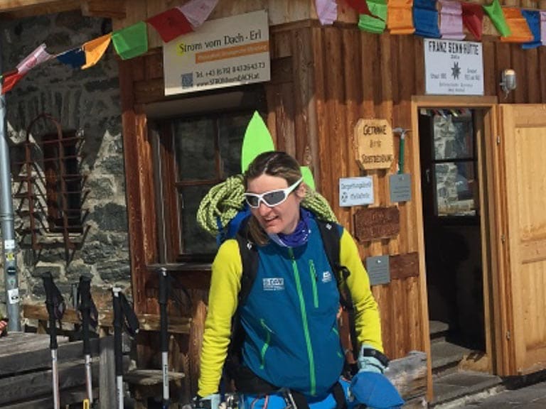 An einer alpinen Berghütte mit Natursteinwänden schnallt Bergwanderführerin Karin Toni Bigel ihre Tourenskier ab.