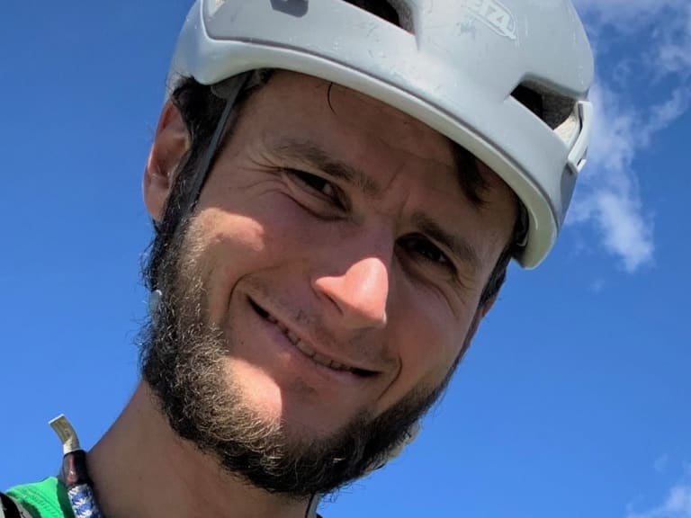 Portrait von Bergführer Jonas Wolkenstörfer mit weißem Kletterhelm und grünem Shirt vor blauem Himmel.