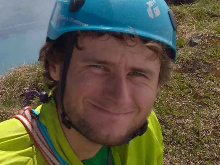 Auf exponiertem Berghang steht Bergführer Jonas Wolkenstörfer mit blauem Helm und türkisfarbenem See im Hintergrund.