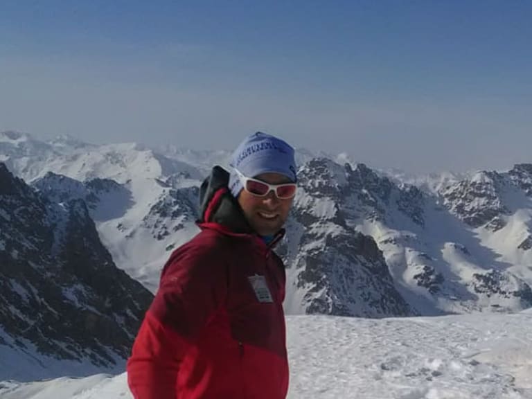 Auf einem schneereichen Gipfel posiert Bergführer Rolando Varesco in roter Jacke für ein Gipfelfoto vor Bergpanorama.