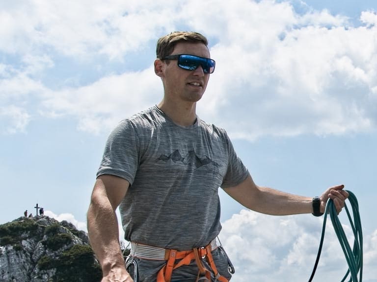 Bergführer Matthias Hahn steht mit Kletterseil und orangem Klettergurt neben einem Holzkreuz auf felsigem Gipfelplateau.