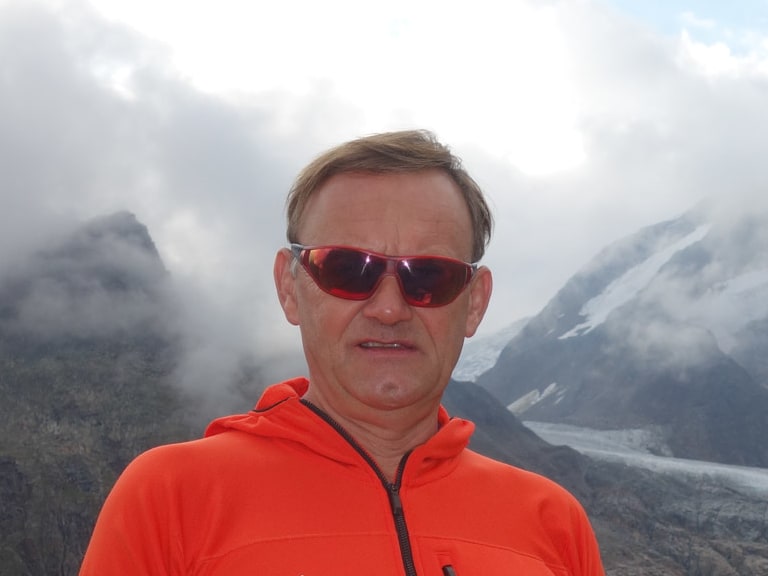 Portrait von Skilehrer Helmut Schramm in orange Softshell-Jacke mit roter Sonnenbrille vor einer Gletscherlandschaft.