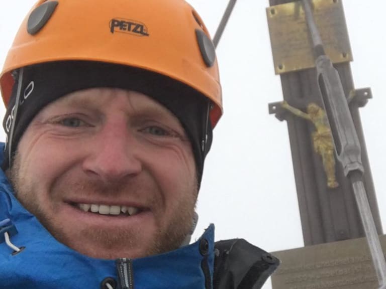 Bergführer Michael Schmidt lächelt am Gipfel mit orangem Petzl-Kletterhelm und blauer Funktionsjacke vor Gipfelkreuz.