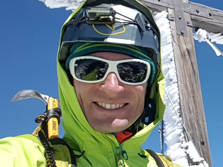Bergführer Martin Riener steht mit zweiter Person vor verreiftem Holzkreuz auf Berggipfel bei klarem Winterwetter.
