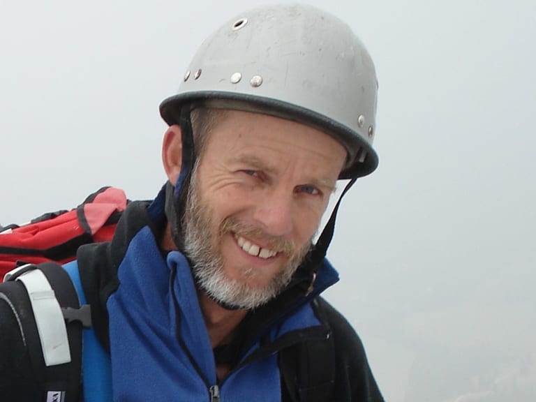 Bergwanderführer Helmut Meyer trägt grauen Kletterhelm und blaue Softshell-Jacke vor schneebedeckten Bergen.