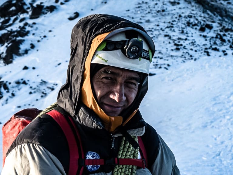Bergführer Marcelo Puruncajas trägt helle Daunenjacke mit weißem Helm und grünem Kletterseil in schneebedeckter Berglandschaft.