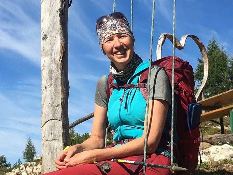 Bergwanderführerin Ruth Zeller sitzt lächelnd auf einer Holzbank am Rastplatz einer Bergwiese mit rotem Wanderrucksack.