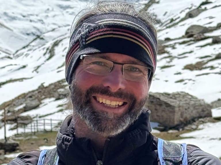 Bergführer Thomas Scherzer mit Brille und gestreiftem Stirnband lächelt vor verschneiter Berglandschaft.