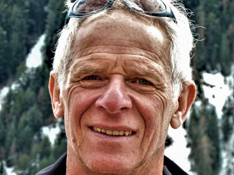 Im Winter zeigt ein Portrait Bergführer Hermann Böhler mit Skibrille auf der Stirn vor verschneiten Nadelbäumen.