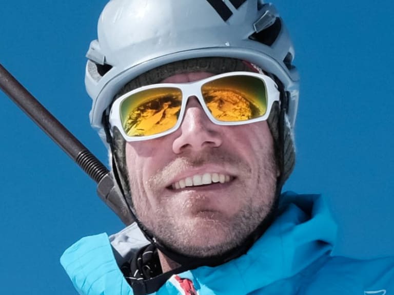 Bergführer Michael Eberharter lächelt in die Kamera, trägt weißen Helm und türkis-blaue Skijacke am Gipfel.