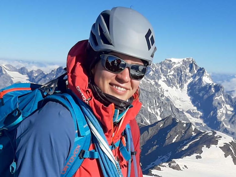 Bergwanderführerin Tatjana Reichert steht mit weißem Helm und roter Jacke auf verschneitem Gipfel vor Hochgebirgspanorama.