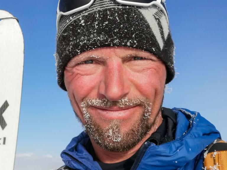 Bergführer Gerald Stranzinger trägt schwarze Mütze und weiße Skibrille auf der Stirn vor blauem Himmel mit Wolkendecke.