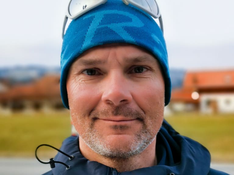 Portrait von Bergführer Tadej Zorman mit blauer Strickmütze und dunkler Funktionsjacke vor bewaldeten Hügeln und Gebäuden mit orangenen Dächern.