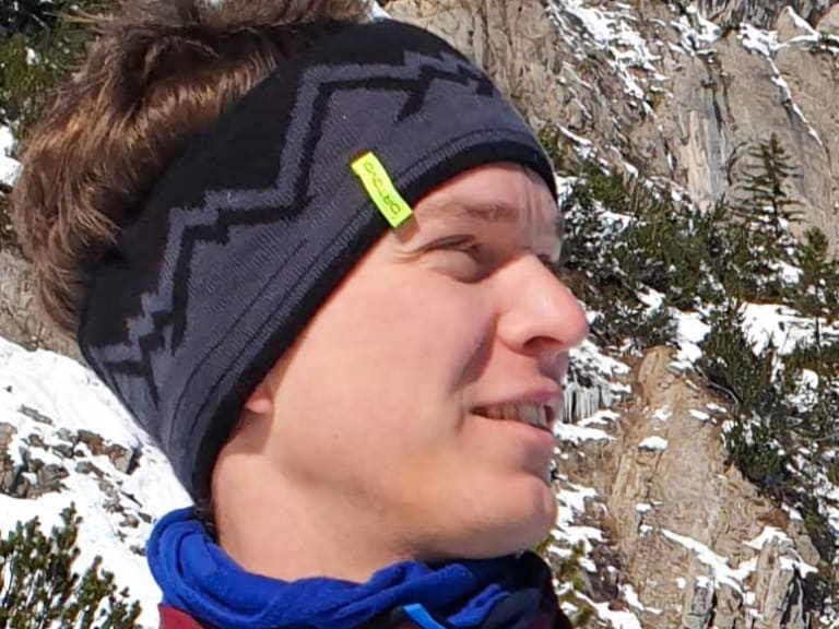 Bergwanderführer Uwe Roth macht ein Selfie vor verschneiten Felswänden und trägt blaue Outdoorjacke mit Alpine Welten Logo.