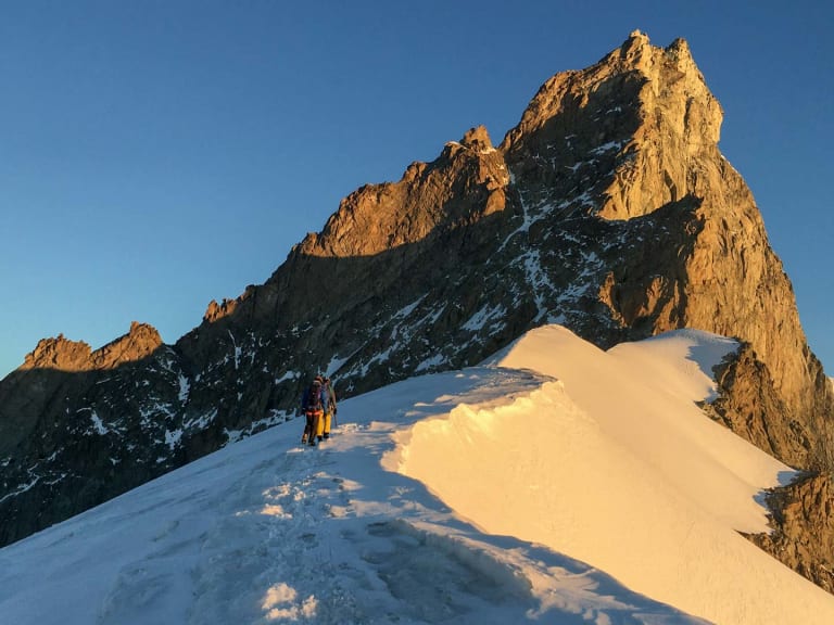 Zwei Bergsteiger in Seilschaft bewegen sich auf schmalem Firnkamm aufwärts, Bergführer Davide Marinovich führt im Morgenlicht zum Felsgipfel.