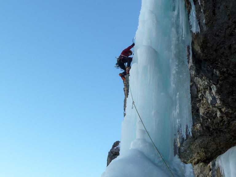 An einer steilen Eiswand klettert Bergführer Davide Marinovich in roter Winterjacke mit Helm und Eisgeräten gesichert am Seil.