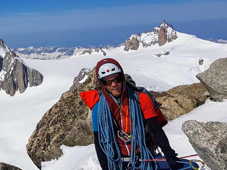 Auf einem schneebedeckten Felsvorsprung steht Bergwanderführer Christoph Knappe in roter Bergbekleidung mit weißem Helm und blauem Seil.