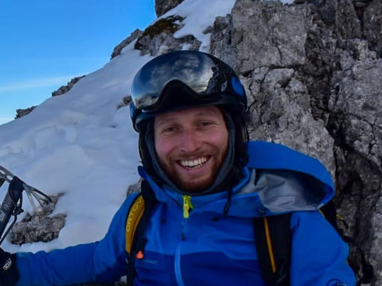 Bergwanderführer Bas Pijpers sitzt lächelnd in blauer Winterjacke mit Eispickel auf verschneitem Felsgrat.