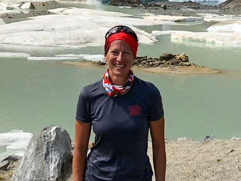 Vor einem Gletschersee mit Eisschollen steht Bergwanderführerin Alexandra Querl in dunkler Funktionskleidung und rotem Stirnband.