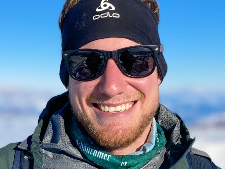 Bergführer Simon Deckenbrock lächelt mit Sonnenbrille und grüner Jacke vor schneebedeckten Berggipfeln.