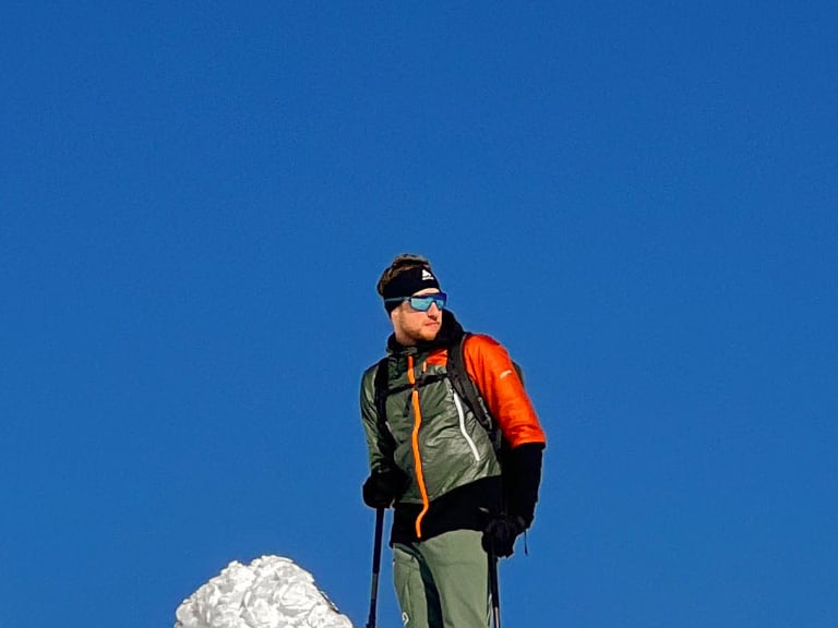 Bergführer Simon Deckenbrock steht auf Tourenski in orange-schwarzer Jacke vor intensiv blauem Himmel.