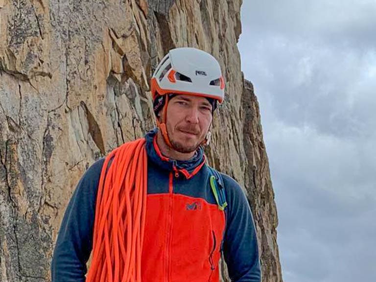 An einer senkrechten Felswand steht Bergführer Márk Salamon mit orange Kletterweste und aufgerolltem Seil.