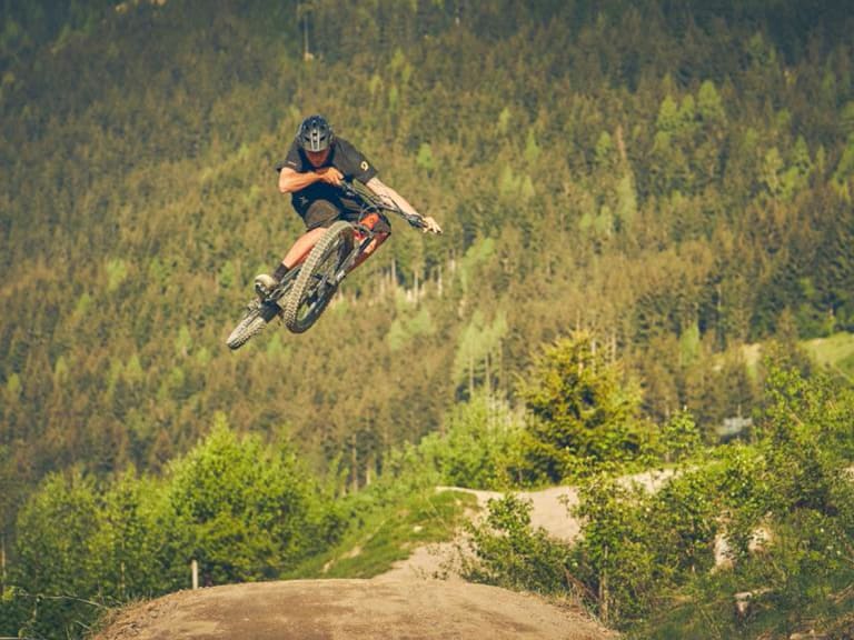 Bergführer Tobias Riedlsperger führt einen Mountainbike-Sprung über eine Erdaufschüttung im bewaldeten Gelände aus.