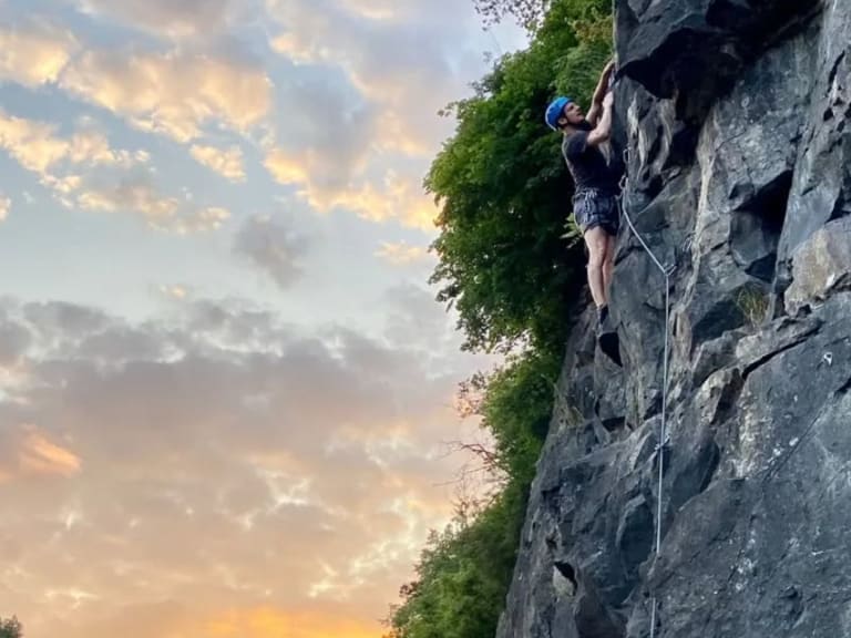 An einer steilen Felswand klettert Bergwanderführer Julian Götz mit blauem Helm und weißem Kletterseil bei Abendstimmung.