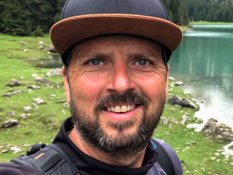 Bergwanderführer Thomas Ziegler macht ein Selfie vor einem türkis-grünen Bergsee mit bewaldeten Hängen im Hintergrund.