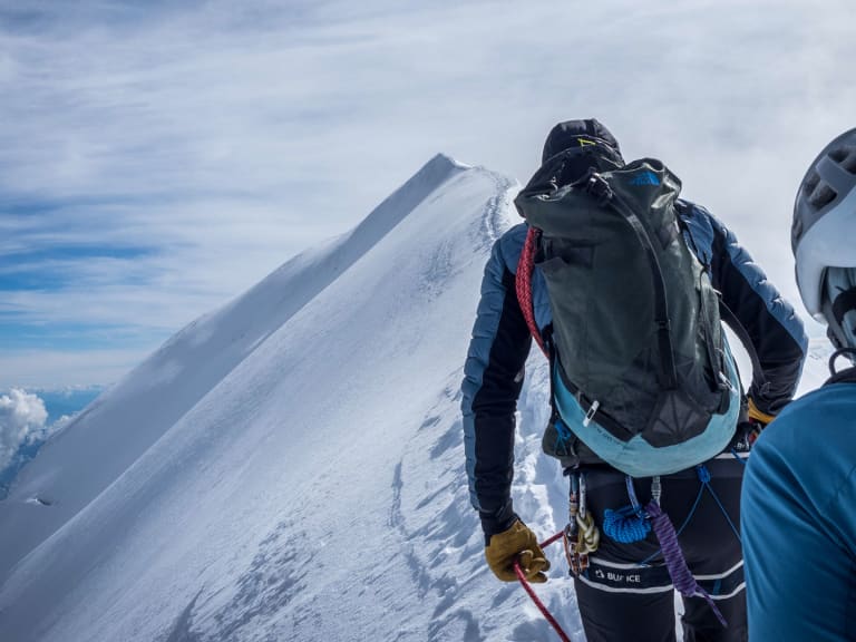 Zwei Bergsteiger bewegen sich angeseilt auf einem schmalen, verschneiten Grat oberhalb der Wolken - Bergführer Enrico Trapazzo führt die Tour.