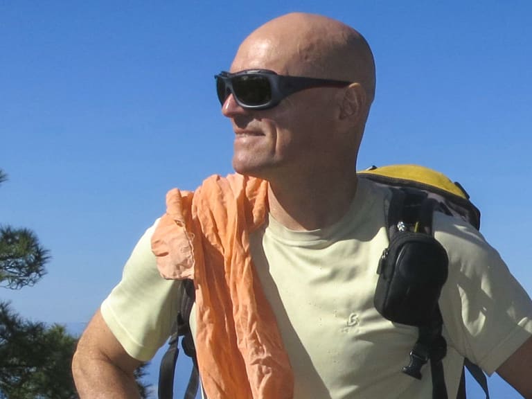 Bergwanderführer Reinald Zölch steht mit Rucksack vor mediterraner Hügellandschaft mit Meer im Hintergrund.