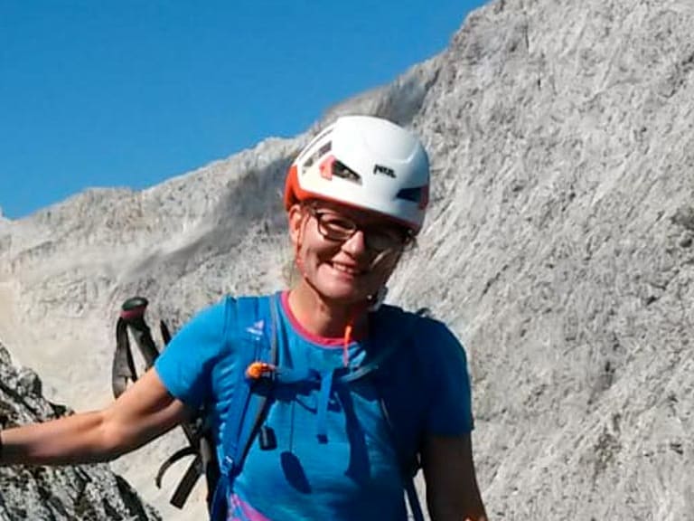 Zwischen hellen Kalksteinfelsen steht Bergwanderführerin Simone Thater mit weißem Helm und Wanderstöcken vor Bergkulisse.