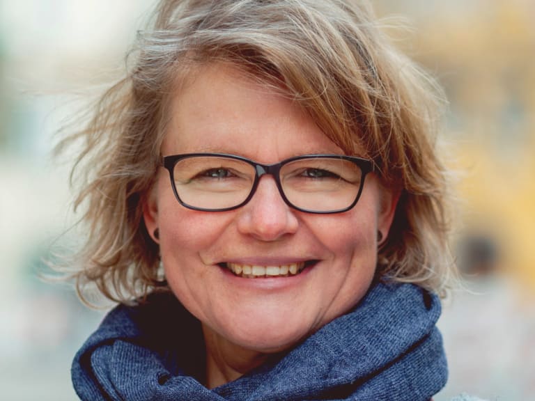 Bergwanderführerin Simone Thater mit blonden Haaren und schwarzer Brille trägt blauen Schal vor städtischer Kulisse.