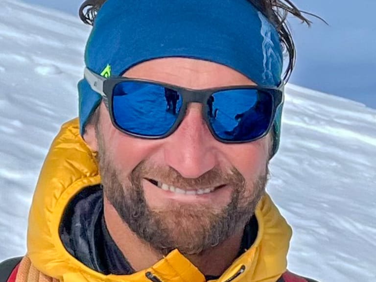 Bergführer Klaus Petz in gelber Daunenjacke mit verspiegelter Sonnenbrille vor verschneiter Berglandschaft bei klarem Sonnenlicht.