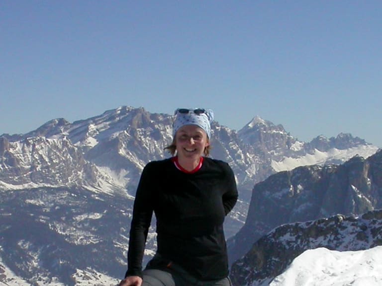 Auf einem verschneiten Gipfel sitzt Bergwanderführerin Birgit Eder mit weißer Mütze vor schneebedeckten Bergen.