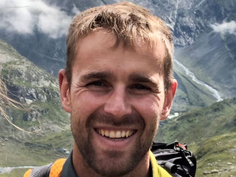 Portrait von Bergwanderführer Benjamin Mäder mit blonden Haaren, gelbem Oberteil und schwarzem Rucksack vor grüner Berglandschaft.