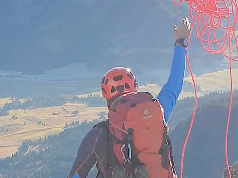 Bergwanderführer Dennis Brida hält rote Seile über dem Kopf und steht mit Klettergurt auf einem Felsvorsprung mit Blick ins Tal.