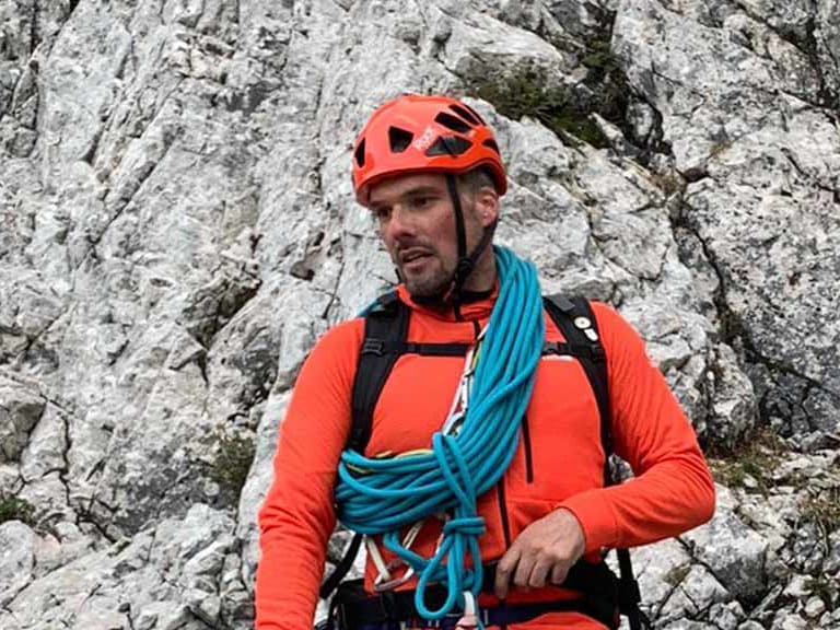 Bergwanderführer Dennis Brida sitzt mit rotem Helm und türkisem Kletterseil auf hellem Kalksteinfels in alpinem Gelände.