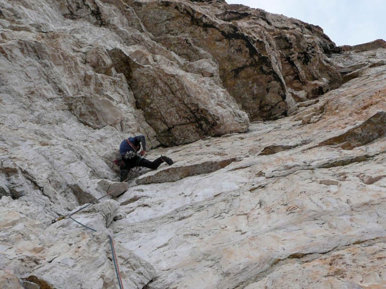 An einer hellen Kalksteinwand klettert Bergführer Klaus Karlstätter mit Helm und orangefarbenem Kletterseil zwischen Felsspalten.