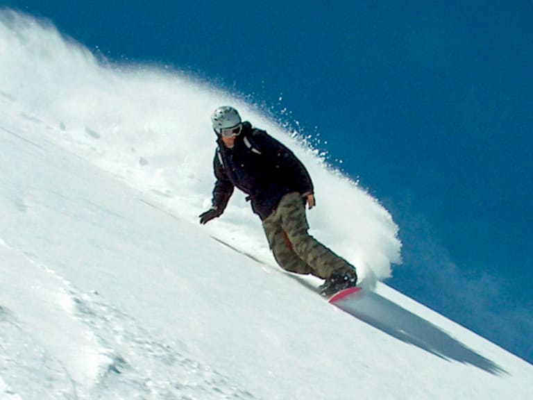 Bergführer Markus Arzberger fährt auf einem Snowboard einen steilen Schneehang hinab und wirbelt dabei Pulverschnee auf.