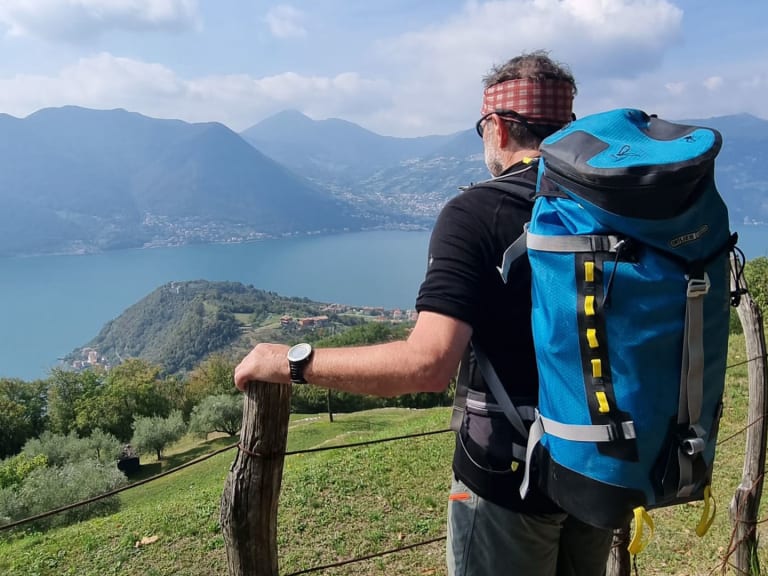 Bergwanderführer Horst Kopelent mit rot-weiß kariertem Stirnband und blauem Rucksack blickt über einen großen Alpensee.