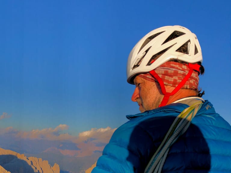 Bergwanderführer Horst Kopelent mit weißem Helm und rotem Tuch blickt zu den Dolomiten-Felswänden bei goldener Abendsonne.