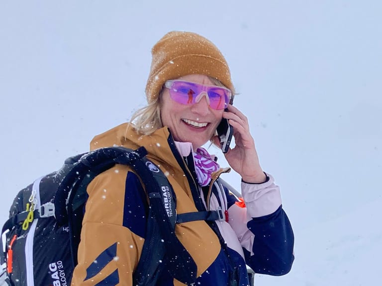 In blau-oranger Winterkleidung telefoniert Bergwanderführerin Stefanie Erdmann neben Holzschild im Schnee.