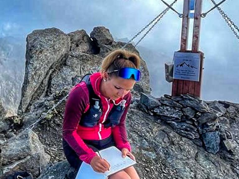Bergwanderführerin Alina Elisabeth Leitinger schreibt in das Gipfelbuch auf felsigem Gipfel neben einem Holzkreuz.
