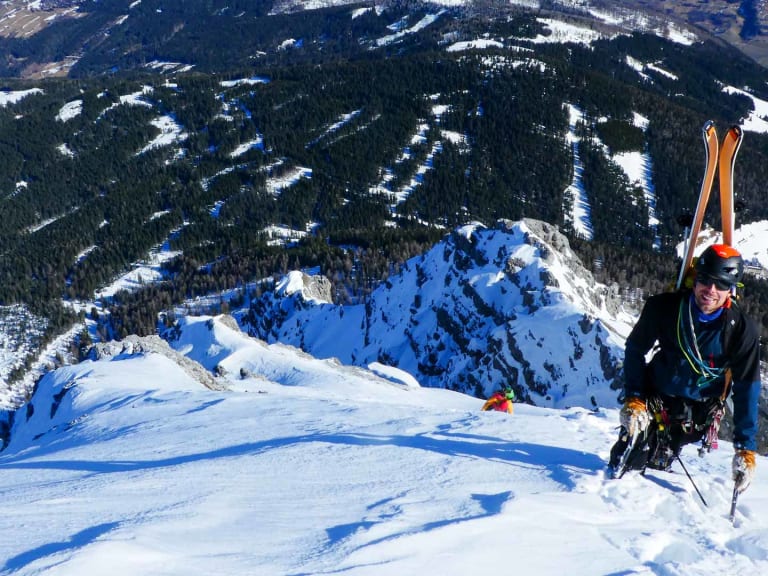 Auf einem verschneiten Gipfel steht Bergführer Lukas Seedoch mit orange Skiern auf dem Rücken befestigt.