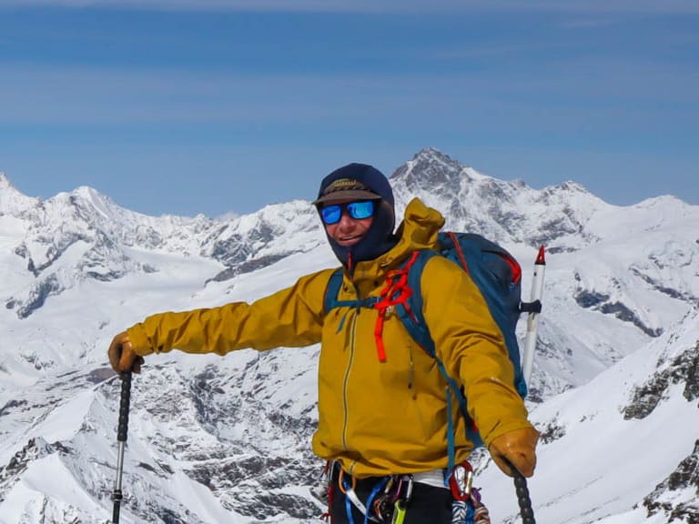 In gelber Winterjacke und mit Steigeisen steht Bergführer Jakob Fink auf verschneitem Hang vor Bergpanorama.