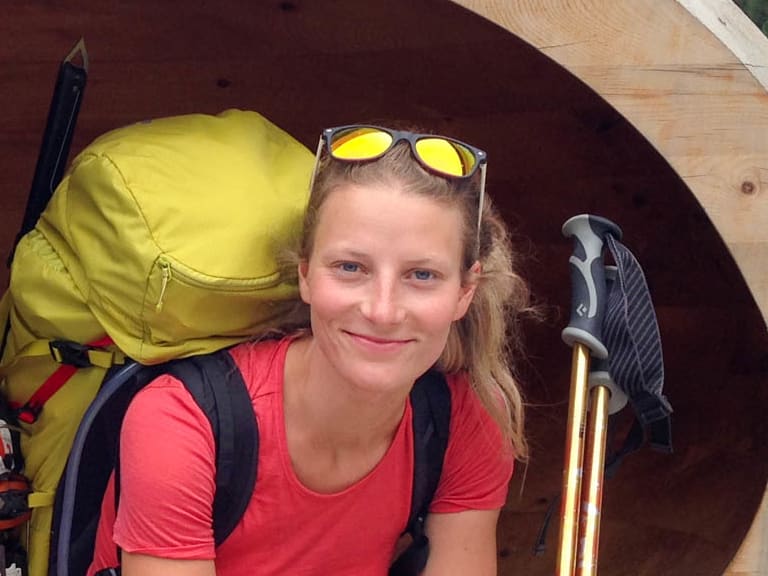 In einer hellen Holzröhre sitzt Bergwanderführerin Anna Biller mit gelbem Trekkingrucksack und blickt in die Kamera.