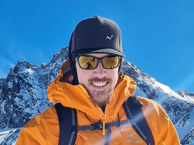 Bergwanderführer Timo Reichert lächelt mit Skistöcken in oranger Jacke vor verschneiter Gletscherlandschaft.