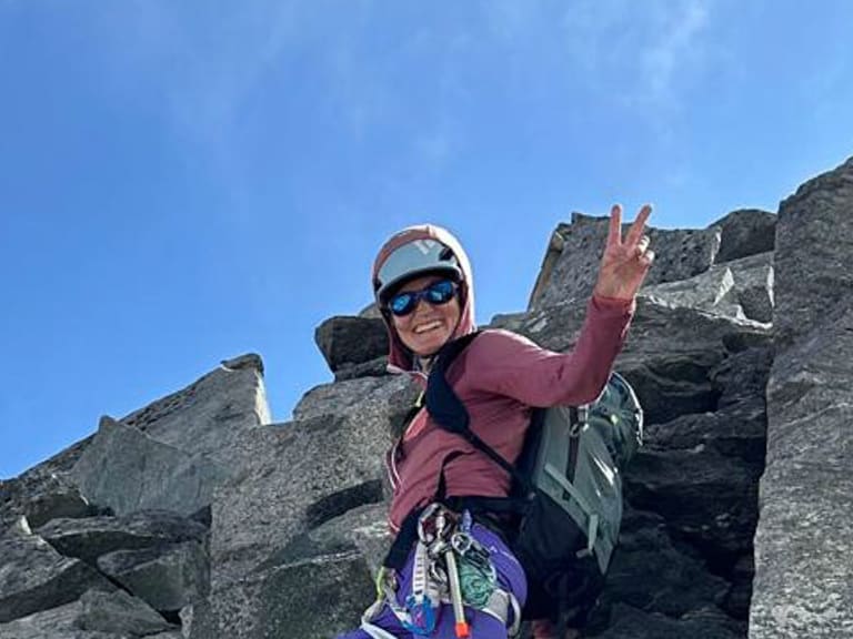 Mit weißem Helm und Klettergurt steht Bergführerin Elisabeth Seibt auf strukturiertem Fels und zeigt ein Victory-Zeichen gegen blauen Himmel.